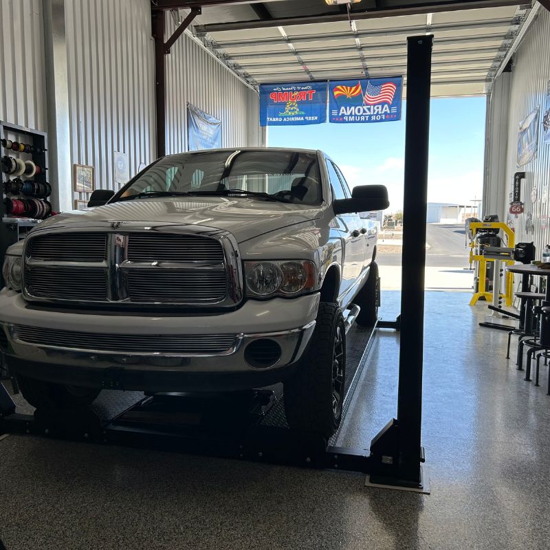 White Dodge 2500 Quad Cab long-bed truck parked on a car lift in a well-lit garage with tools and banners visible in the background.